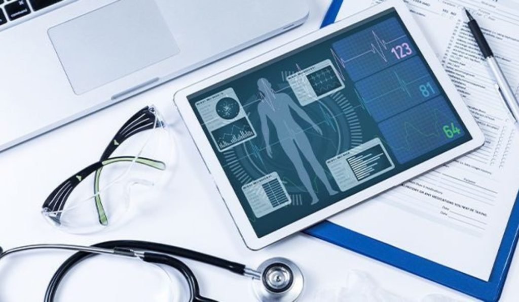 Medical tablet displaying health data with heart rate and blood pressure graphs, surrounded by a stethoscope, glasses, and papers on a desk.
