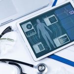 Medical tablet displaying health data with heart rate and blood pressure graphs, surrounded by a stethoscope, glasses, and papers on a desk.