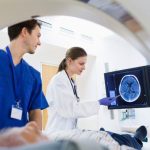 A medical professional and technician reviewing a brain scan on a monitor while a patient lies on a medical examination table.