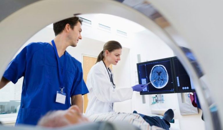 A medical professional and technician reviewing a brain scan on a monitor while a patient lies on a medical examination table.