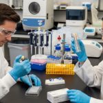 Lab scientists wearing safety goggles and gloves conducting a test with a pipette and sample tray in a pharmaceutical laboratory.