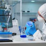 Laboratory technician in protective suit and mask examining a petri dish in a sterile lab with test tubes and lab equipment.