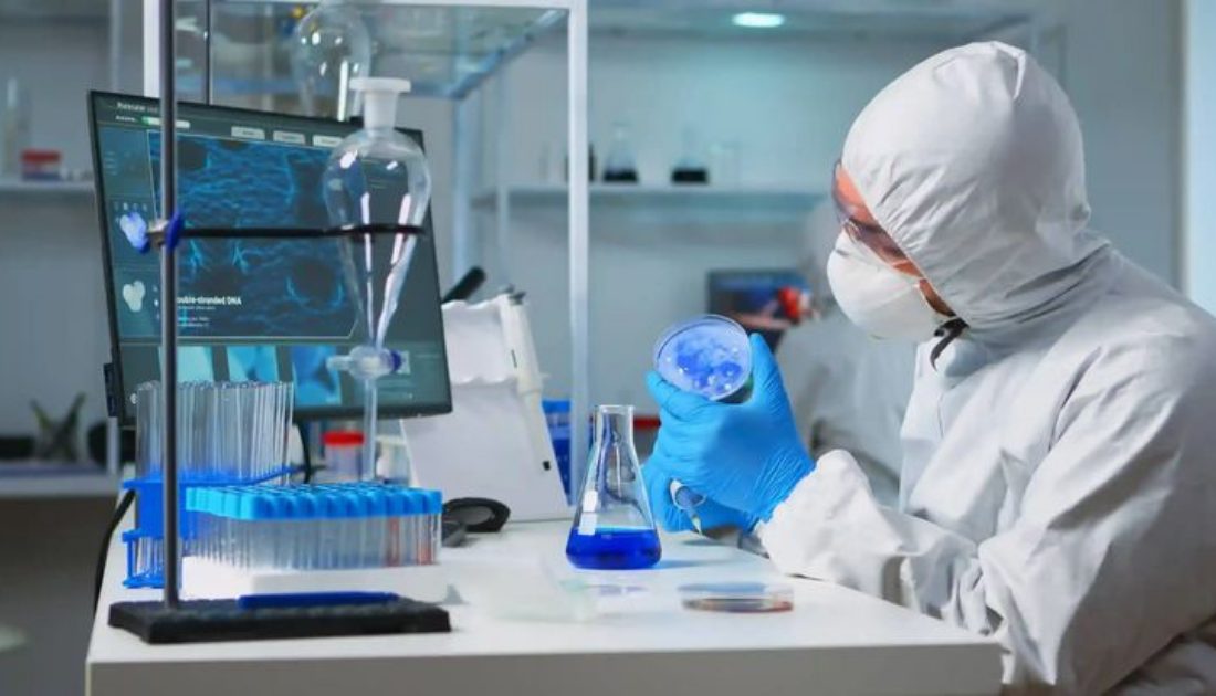 Laboratory technician in protective suit and mask examining a petri dish in a sterile lab with test tubes and lab equipment.