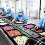 Factory workers in masks and protective uniforms inspecting and sorting cosmetic products on a production line in a clean manufacturing facility.