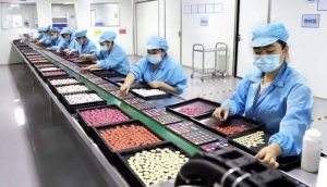 Factory workers in masks and protective uniforms inspecting and sorting cosmetic products on a production line in a clean manufacturing facility.
