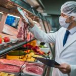 Food safety inspector in a lab coat and hairnet checking meat temperature with a probe in a refrigerated display while holding a clipboard.