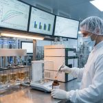 A male QC analyst in a lab coat and mask uses forceps to sample a softgel at a stainless steel workstation featuring a dissolution tester, analytical balance, and monitors with data charts.