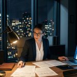 A pharmaceutical professional in a high-rise office at night, intensely reviewing compliance documents under a desk lamp with a glowing laptop dashboard and city skyline in the background.